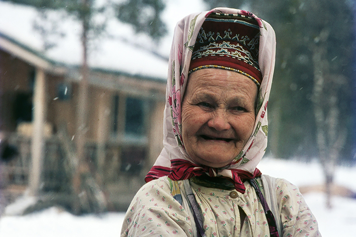Värillinen valokuva hymyilevästä saamelaisesta vanhasta naisesta, jolla on kansallispuku. Taustalla on lumista metsää ja puinen mökki.