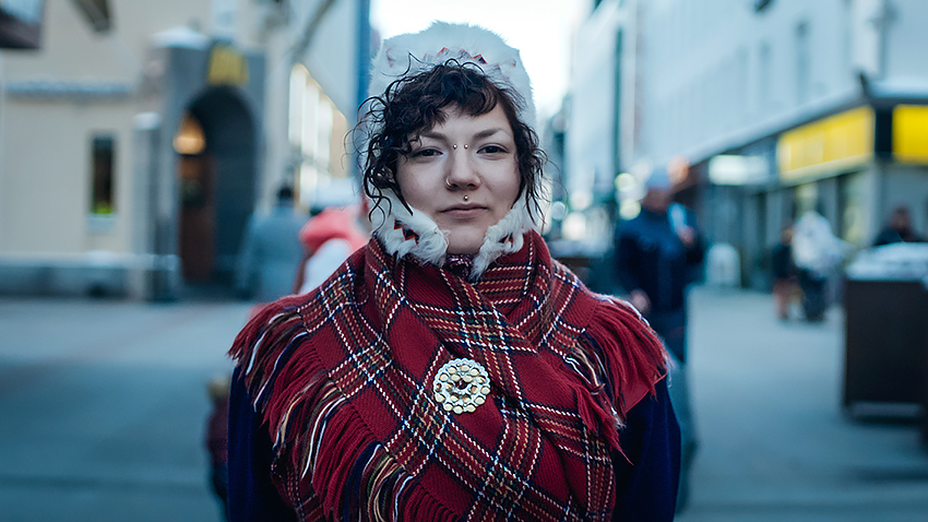 Värillinen valokuva saamelaisesta henkilöstä, joka katsoo suoraan kameraan. Taustalla on vilkas kaupungin katu ja rakennuksia.