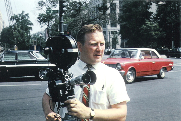 Värillinen valokuva miehestä filmikamera kädessään. Taustalla on vilkas tie, jolla kulkee musta ja punainen vanha auto. Miehellä on valkoinen kauluspaita ja punaraitainen solmio.