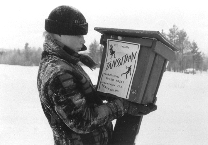 Mustavalkoinen valokuva, jossa talvivaatteinen pipopäinen ihminen kantaa lumisessa maisemassa postilaatikkoa. Postilaatikon kylkeen on liimattu paperilappu, jossa on mainos tansseista.