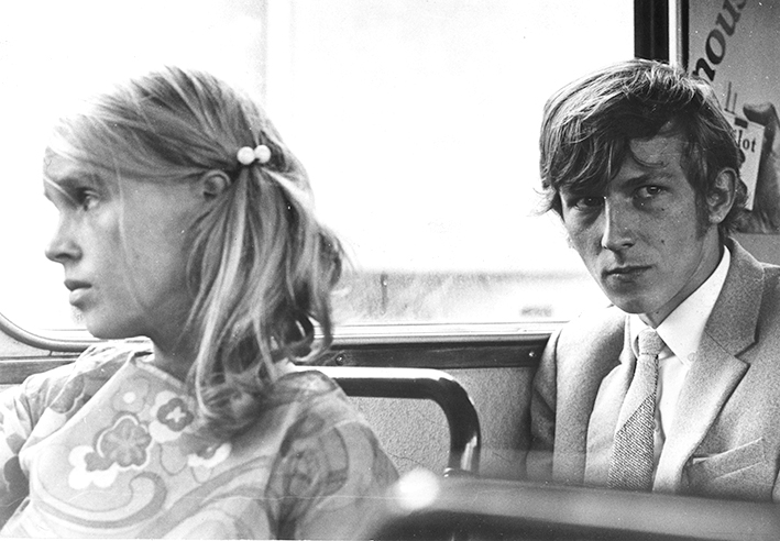 Mustavalkoinen valokuva kahdesta henkilöstä bussissa. Edessä istuvalla on poninhäntä ja kukkapaita, takana istuvalla on solmio ja puvuntakki.