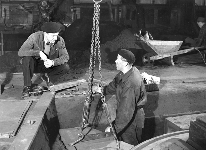 Mustavalkoinen valokuva, jossa kaksi henkilöä keskustelevat. Vasemmanpuoleinen henkilö kyykkää maassa hattu päässään, oikeanpuoleinen seisoo kuopassa työvaatteet päällään ja pitää kiinni roikkuvasta ketjusta. Taustalla on kottikärryt ja rakennustyömaata.